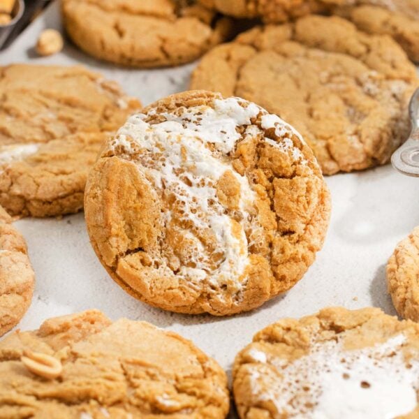 A close-up of a peanut butter cookie with a gooey marshmallow filling oozing out, surrounded by more similar cookies on a light surface.
