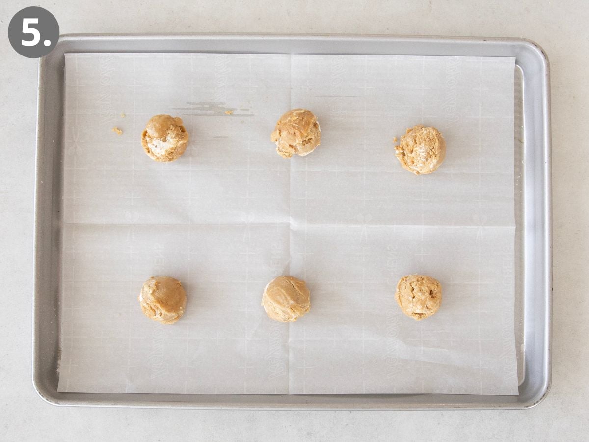 A baking sheet lined with parchment paper holds six evenly spaced scoops of cookie dough, arranged in two rows of three. A gray number 5 is in the top left corner.