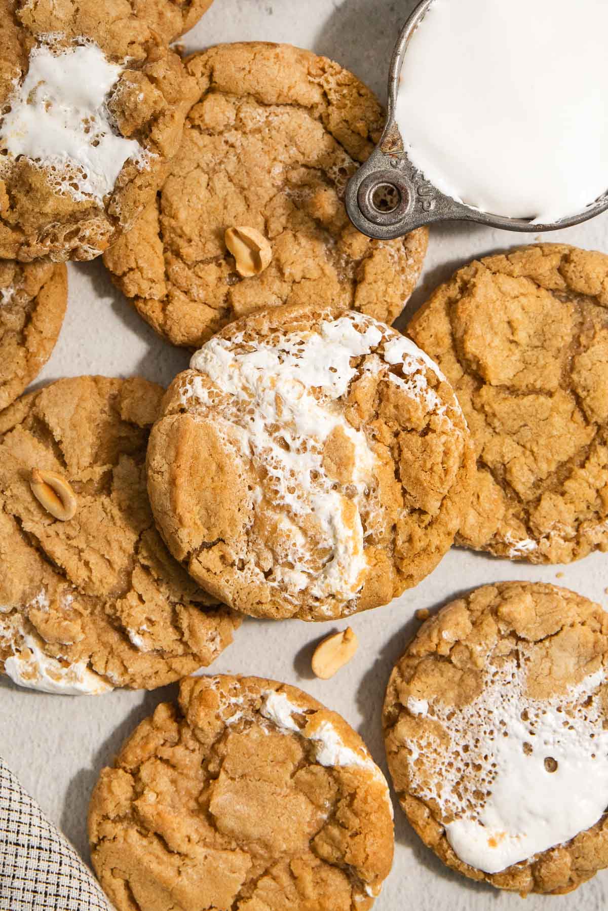 A close-up of several peanut butter cookies with marshmallow swirls, some topped with peanuts. A metal cup filled with milk is partially visible in the top right corner.