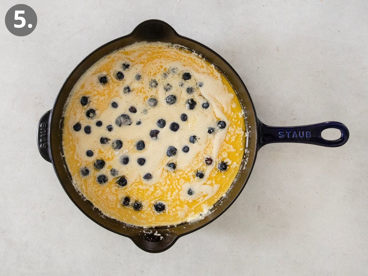A black skillet with a handle sits on a light surface, filled with a yellow batter and scattered blueberries. The skillet is labeled STAUB and the image is marked with a number 5 in the top left corner.