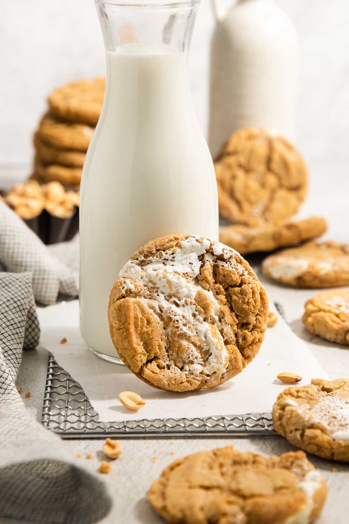 A glass bottle of milk sits on a wire rack with a peanut butter cookie filled with marshmallow cream leaning against it. More cookies and a napkin are scattered around in the background.