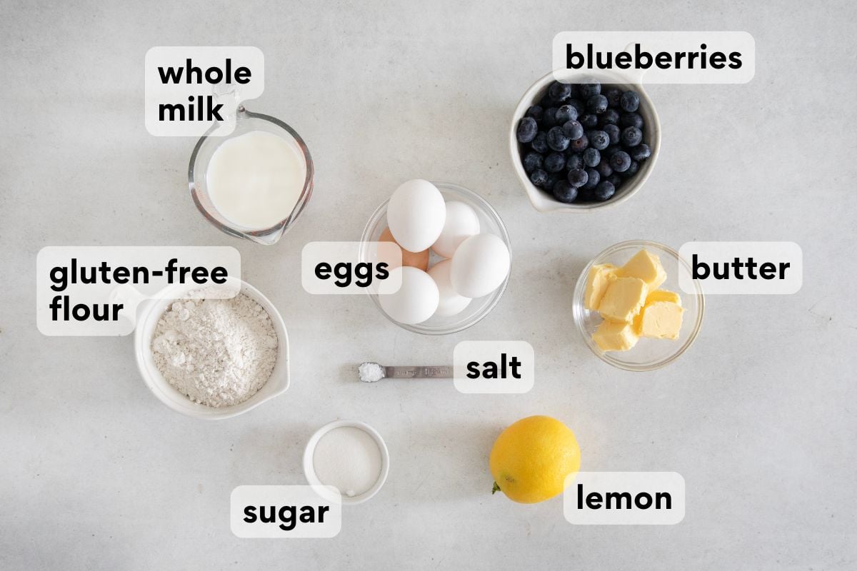 Labeled ingredients on a countertop: whole milk, blueberries, gluten-free flour, eggs, butter, salt, sugar, and a lemon. Each item is in a bowl or cup with clear text labels.