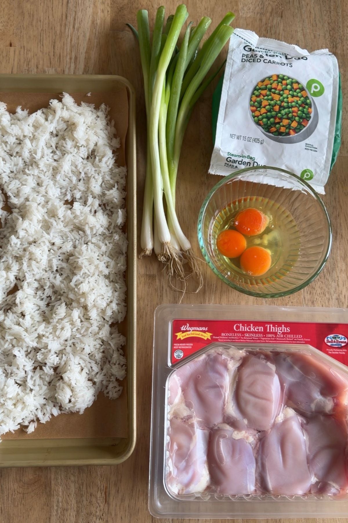 A wooden table with cooked rice on a tray, green onions, a bag of frozen peas and carrots, two cracked eggs in a bowl, and a package of raw chicken thighs.