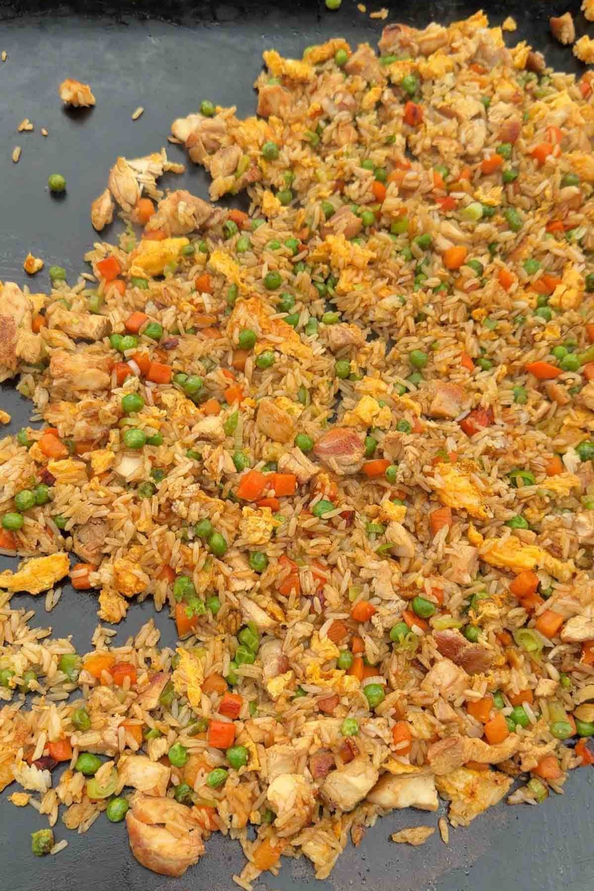 Fried rice with chicken, scrambled eggs, peas, and diced carrots spread on a flat cooking surface.