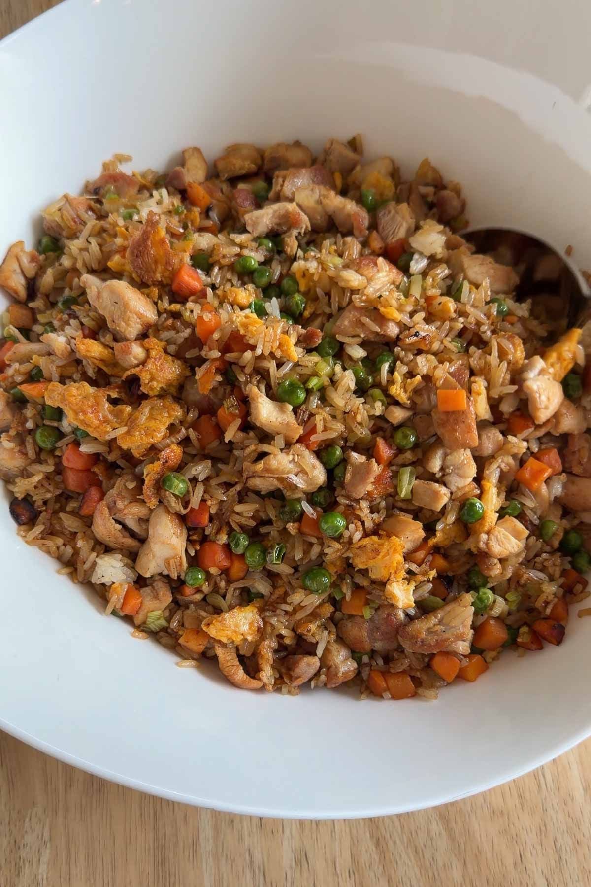 A white bowl filled with fried rice including chopped chicken, peas, diced carrots, green onions, and scrambled egg pieces, with a spoon on the side.