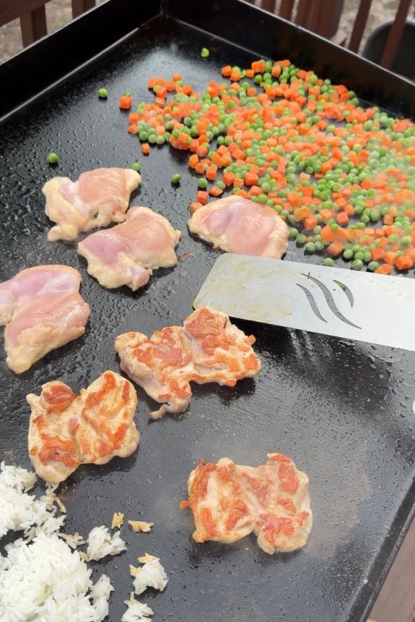Chicken pieces cooking on a flat top grill with a metal spatula, alongside a mix of peas and carrots and some white rice.