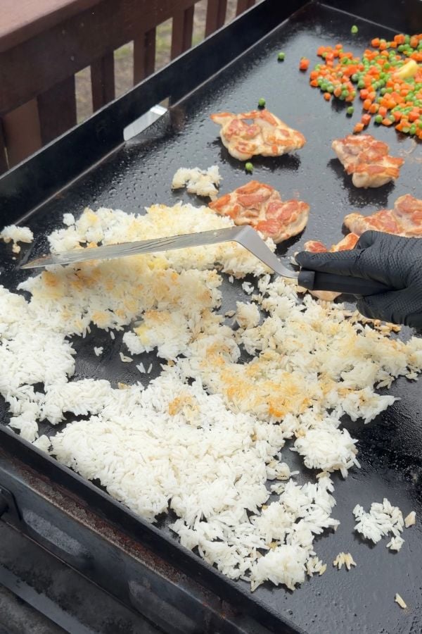 A person wearing a black glove is using a spatula to cook white rice on a flat-top grill, with chicken pieces and mixed vegetables visible in the background.