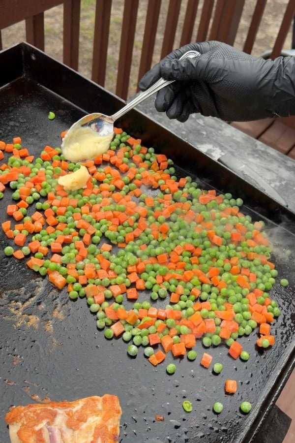 A gloved hand holding a spoon adds a dollop of butter to peas and diced carrots cooking on a flat-top grill outdoors. Steam rises from the vegetables.