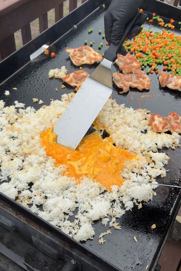 A gloved hand uses a spatula to mix egg with white rice on a griddle, with pieces of meat and a scattered mix of peas and carrots in the background.