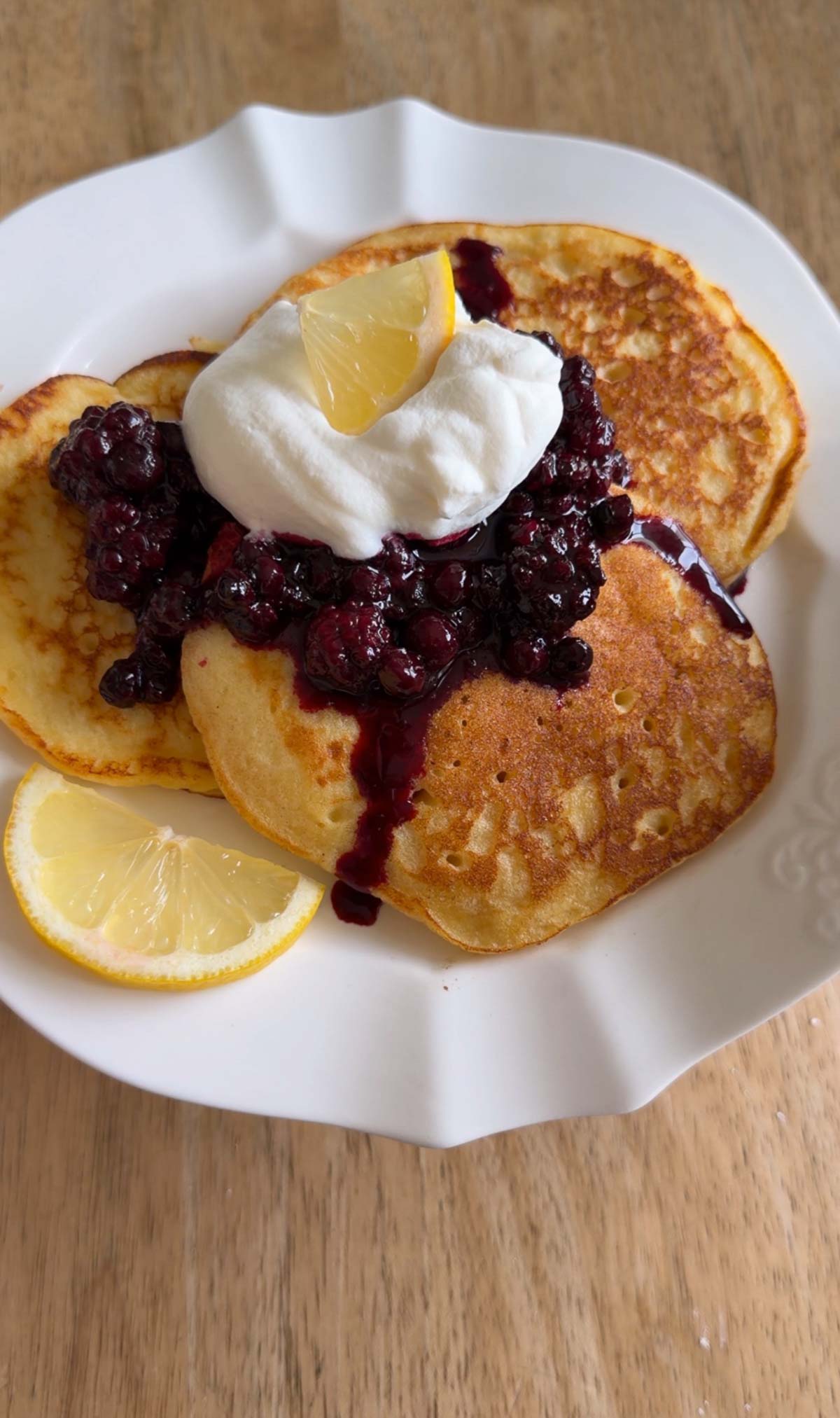 Three pancakes topped with whipped cream, berry compote, and a lemon slice on a white plate, with another lemon slice on the side, set on a wooden table.