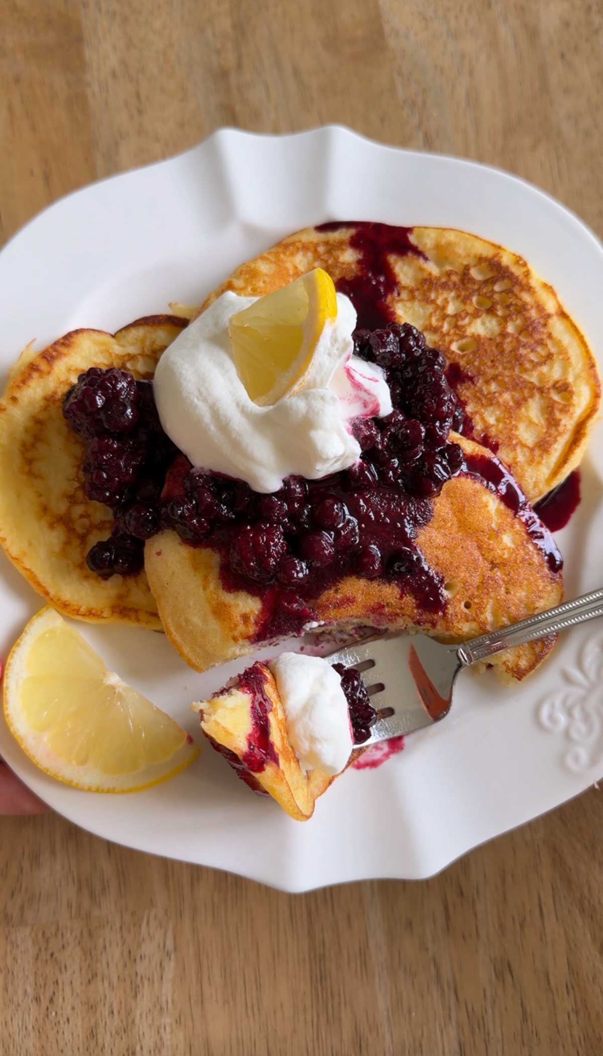 Pancakes topped with blueberry compote, whipped cream, and a lemon wedge on a white plate. A fork holds a bite of pancake with toppings. The plate is set on a wooden surface.