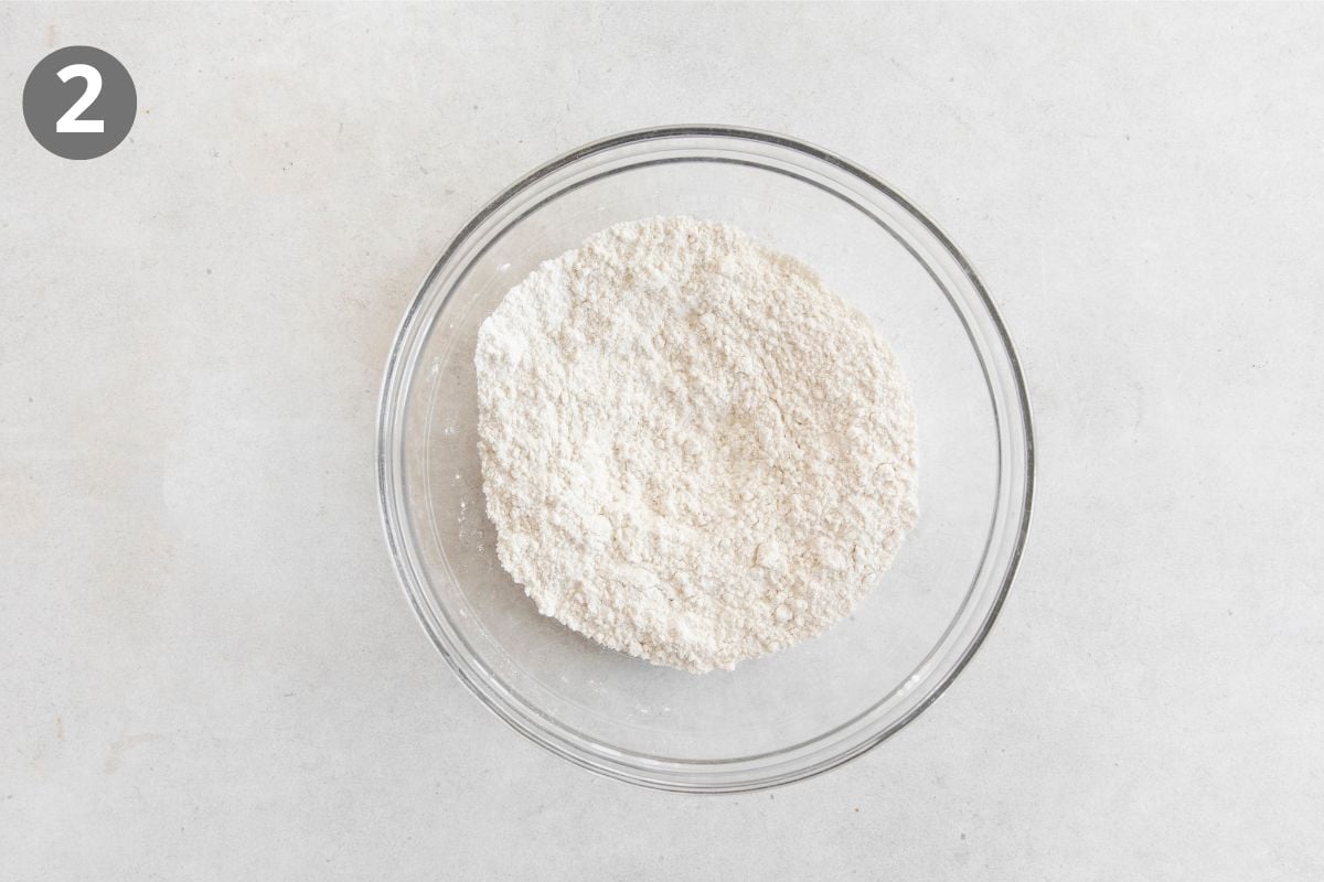 A glass bowl filled with flour sits on a light surface. A gray circle with the number 2 is in the top left corner.