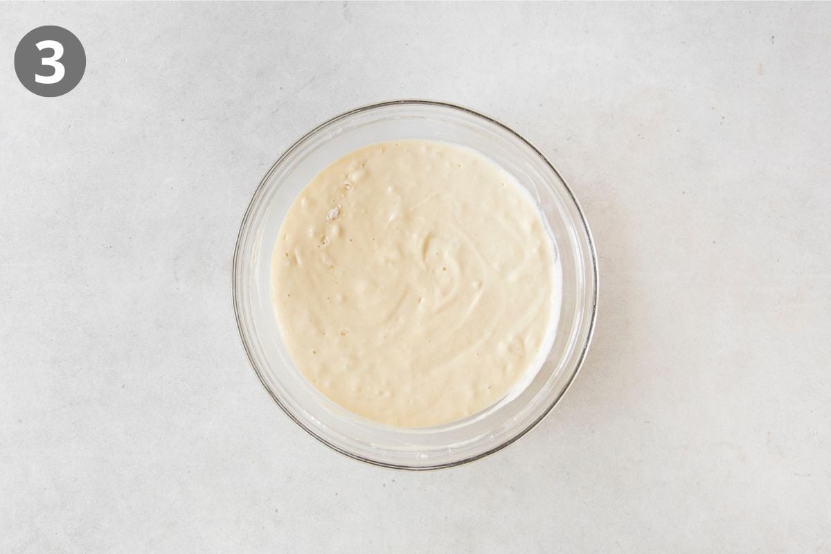 A glass bowl filled with creamy batter sits on a light surface. A number 3 in a dark circle appears in the upper left corner, indicating the third step in a recipe.
