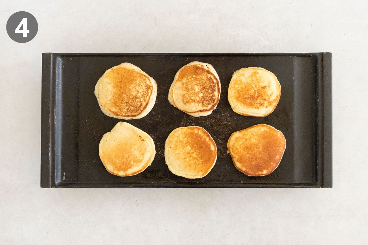 Six golden brown pancakes are cooking on a black griddle, viewed from above. A grey circle with the number 4 is in the top left corner of the image.