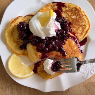 Three pancakes on a white plate topped with whipped cream, berry compote, and a lemon wedge. A fork holds a bite with whipped cream and berries. Another lemon wedge sits on the side.