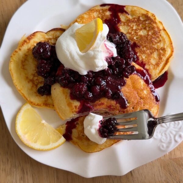 Three pancakes on a white plate topped with whipped cream, berry compote, and a lemon wedge. A fork holds a bite with whipped cream and berries. Another lemon wedge sits on the side.