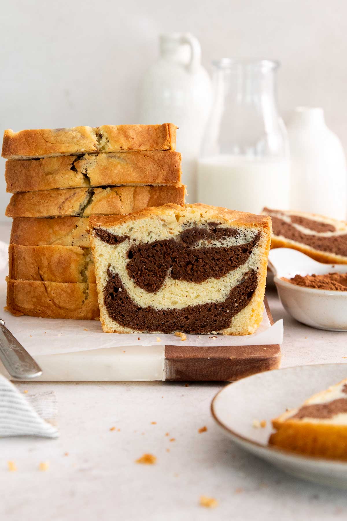 A sliced marble loaf cake is displayed on a cutting board, showing its swirled chocolate and vanilla pattern. Several slices are stacked behind it, with milk jars and bowls in the background.