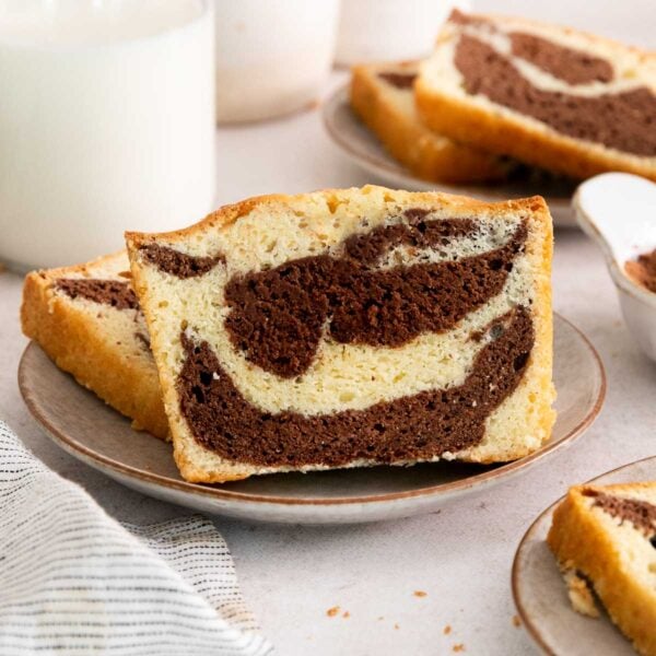 A slice of marble cake with chocolate and vanilla swirls sits on a plate, with more slices and a cup visible in the background.