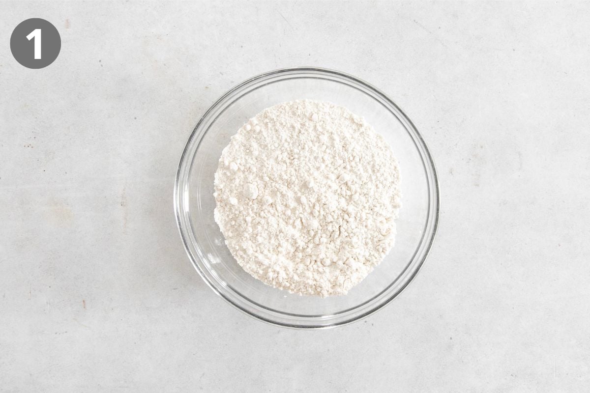 A glass bowl filled with flour sits on a light gray surface. A small gray circle with the number 1 appears in the top left corner of the image.