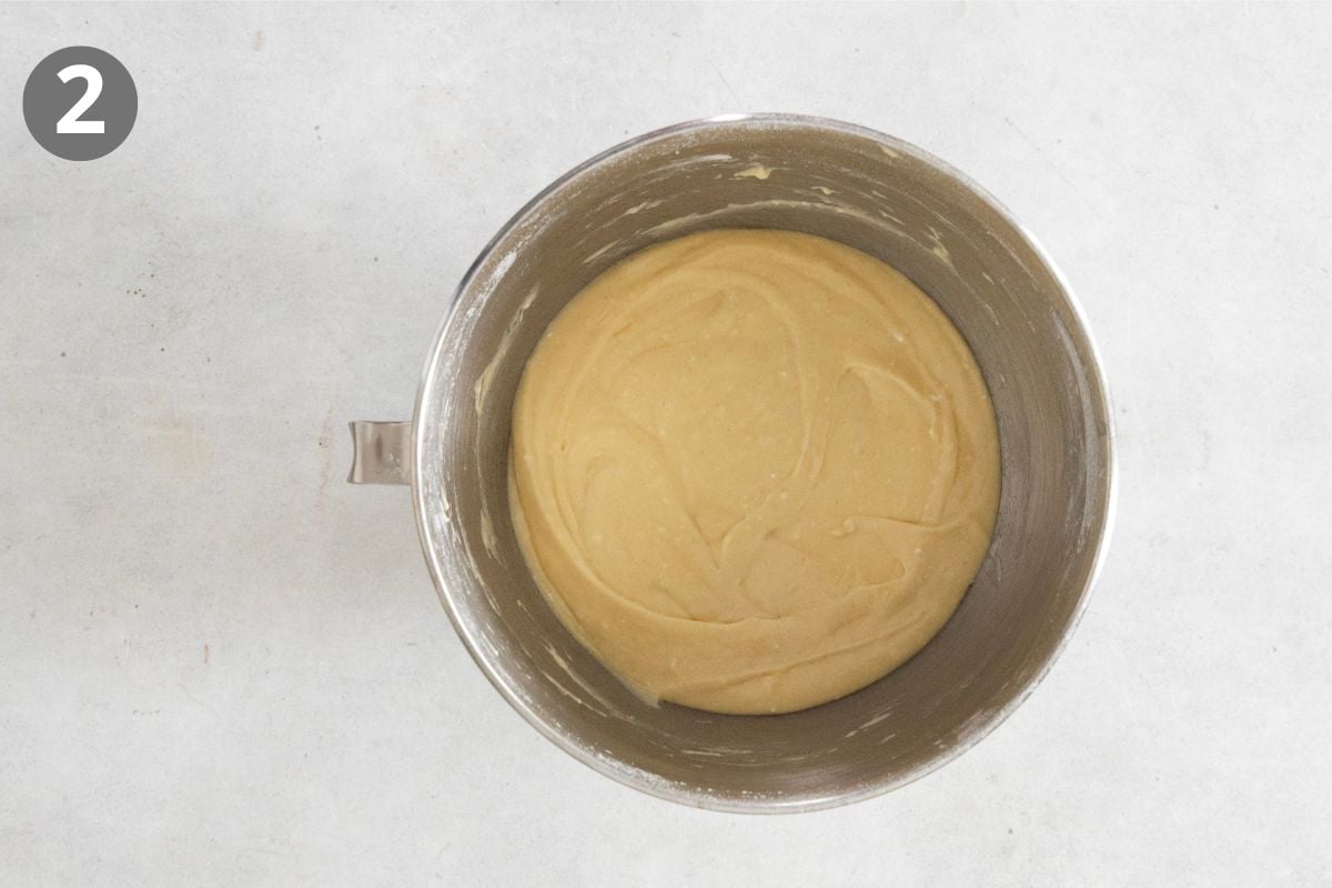A metal mixing bowl with smooth, light yellow cake batter sits on a white surface. A gray circle with the number 2 is in the top left corner.