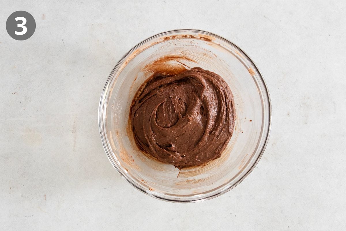 A glass bowl with chocolate batter sits on a light-colored countertop. The number 3 appears in a gray circle in the top left corner.