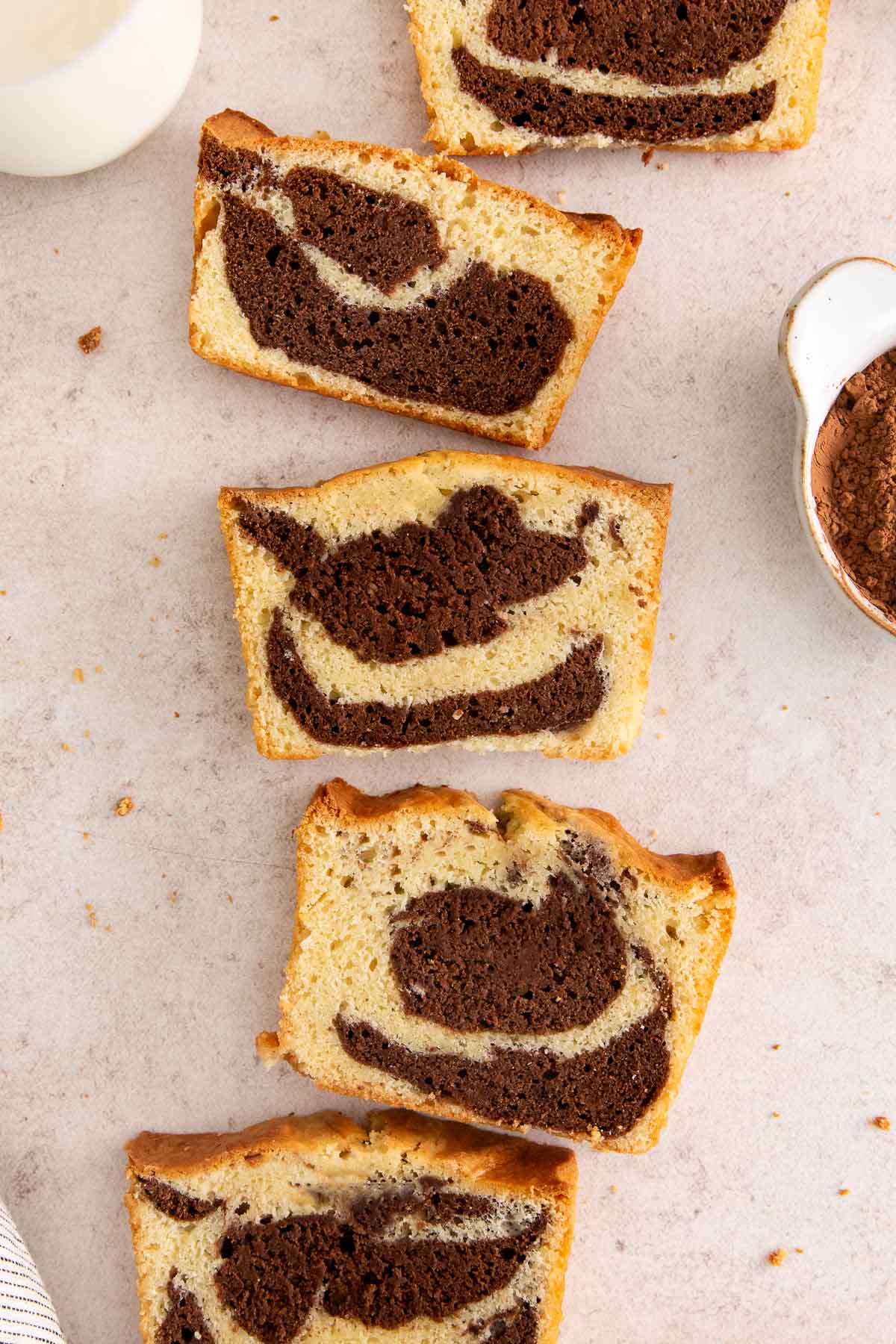 Five slices of marble cake are arranged on a light surface, showing a mix of chocolate and vanilla swirls. A bowl of cocoa powder and part of a milk jug are also visible in the background.
