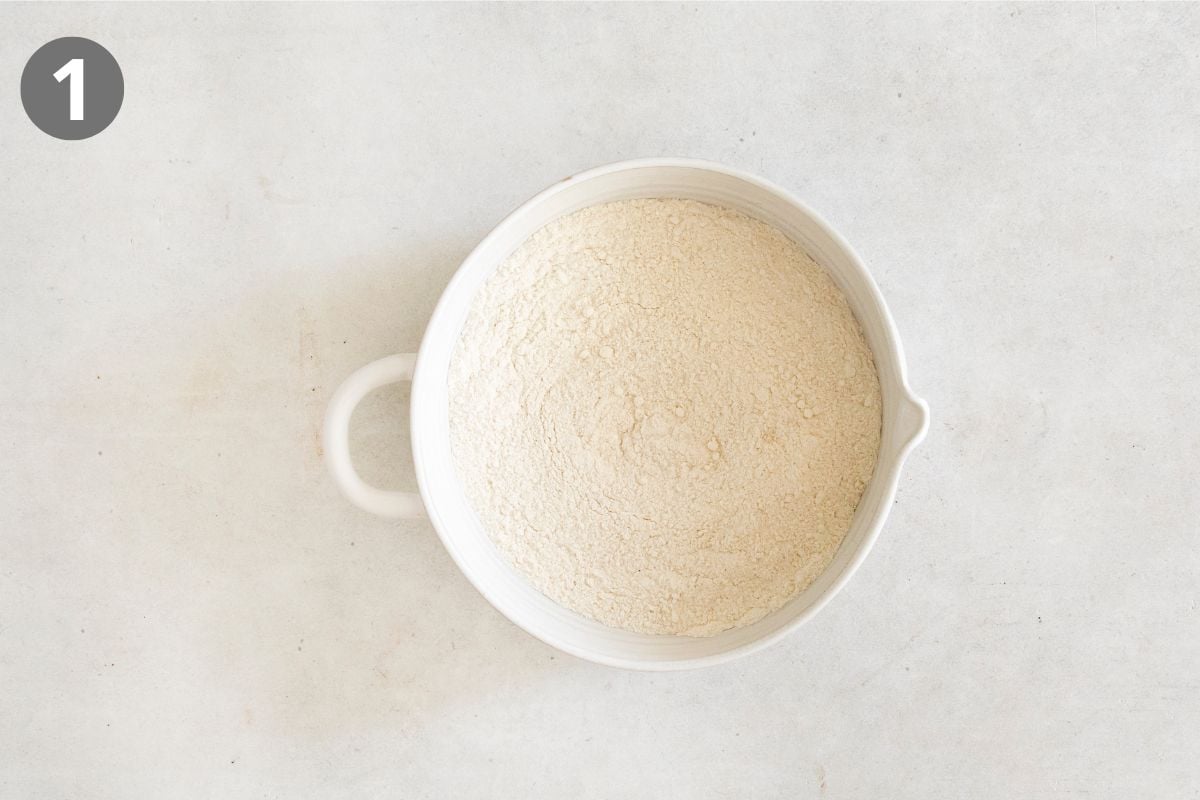 A white mixing bowl filled with flour sits on a light gray surface. A gray circle with the number 1 is in the top left corner.