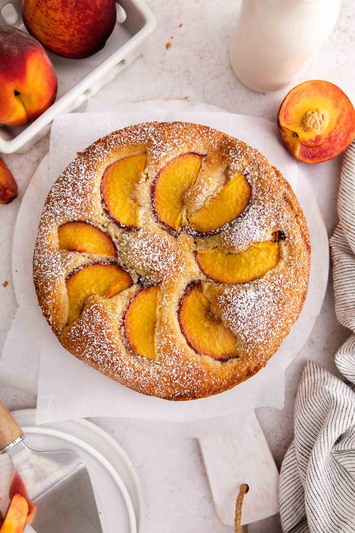 A round peach cake topped with powdered sugar and peach slices sits on parchment paper, surrounded by fresh peaches, a knife, and a beige-striped cloth.