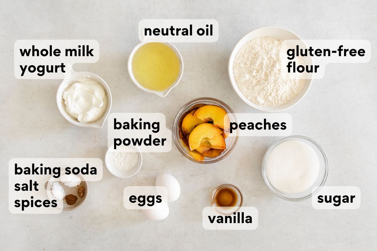 Top-down view of labeled baking ingredients: whole milk yogurt, eggs, peaches, neutral oil, gluten-free flour, sugar, vanilla, baking powder, and baking soda, salt, and spices, arranged on a white surface.
