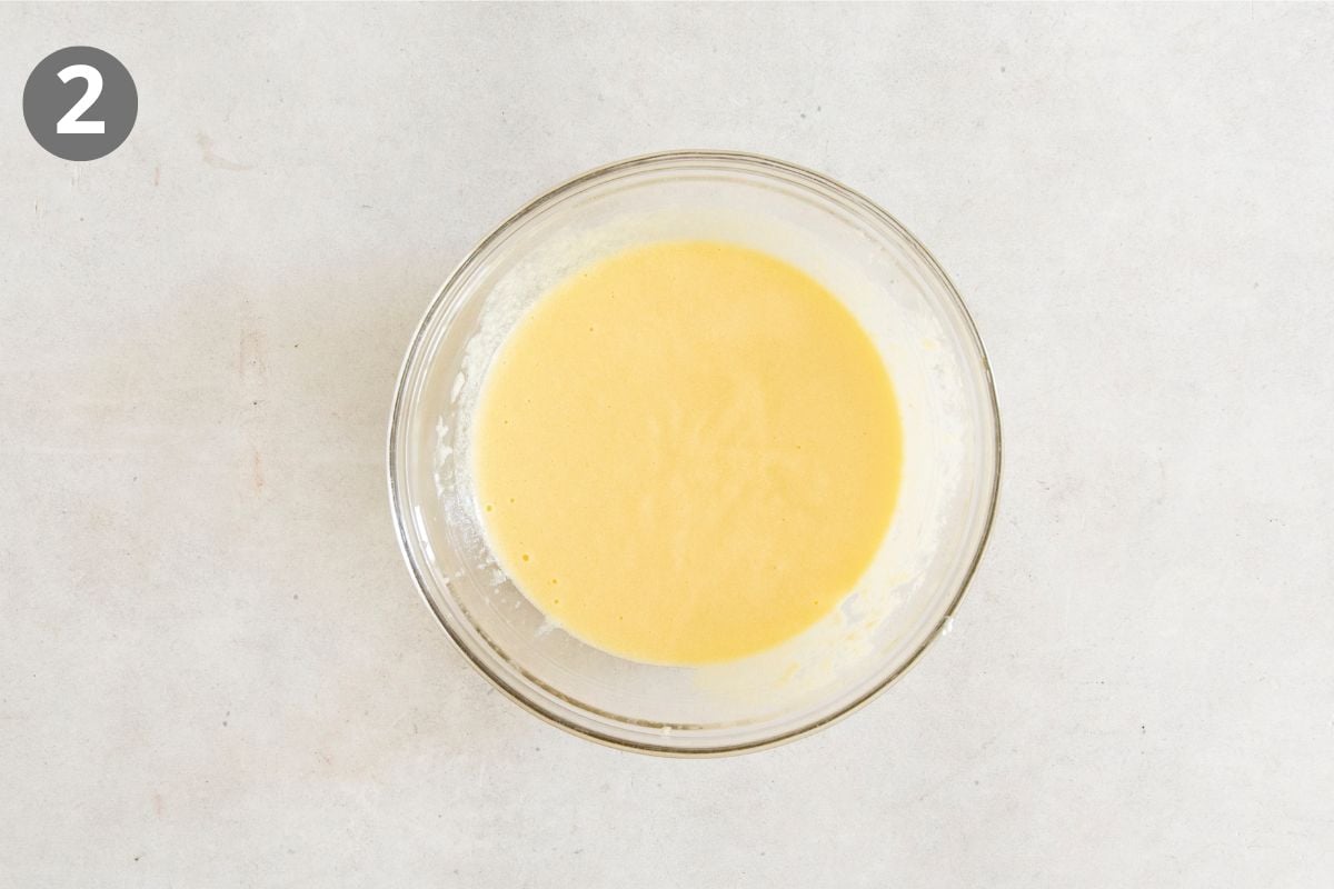 A glass bowl filled with smooth, pale yellow batter sits on a light-colored surface. A gray circle with the number 2 is in the top left corner.