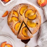 A round peach cake, topped with peach slices and powdered sugar, has one slice being served. Fresh peach wedges and a striped towel are nearby on a light surface.