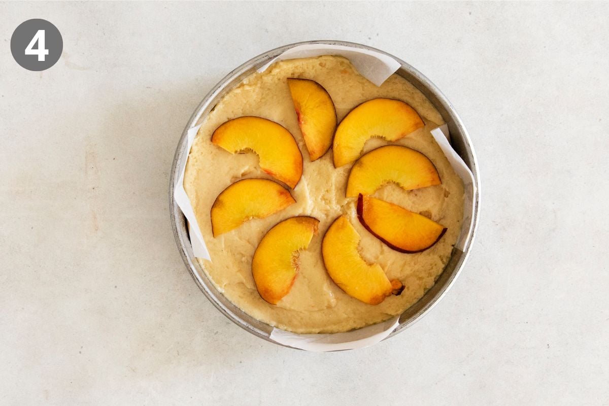 A round baking pan with parchment paper holds cake batter topped with peach slices arranged in a circular pattern. A small number 4 is in the upper left corner.