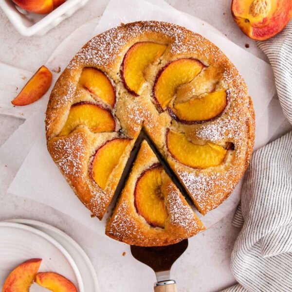 A round peach cake, topped with peach slices and powdered sugar, has one slice being served. Fresh peach wedges and a striped towel are nearby on a light surface.