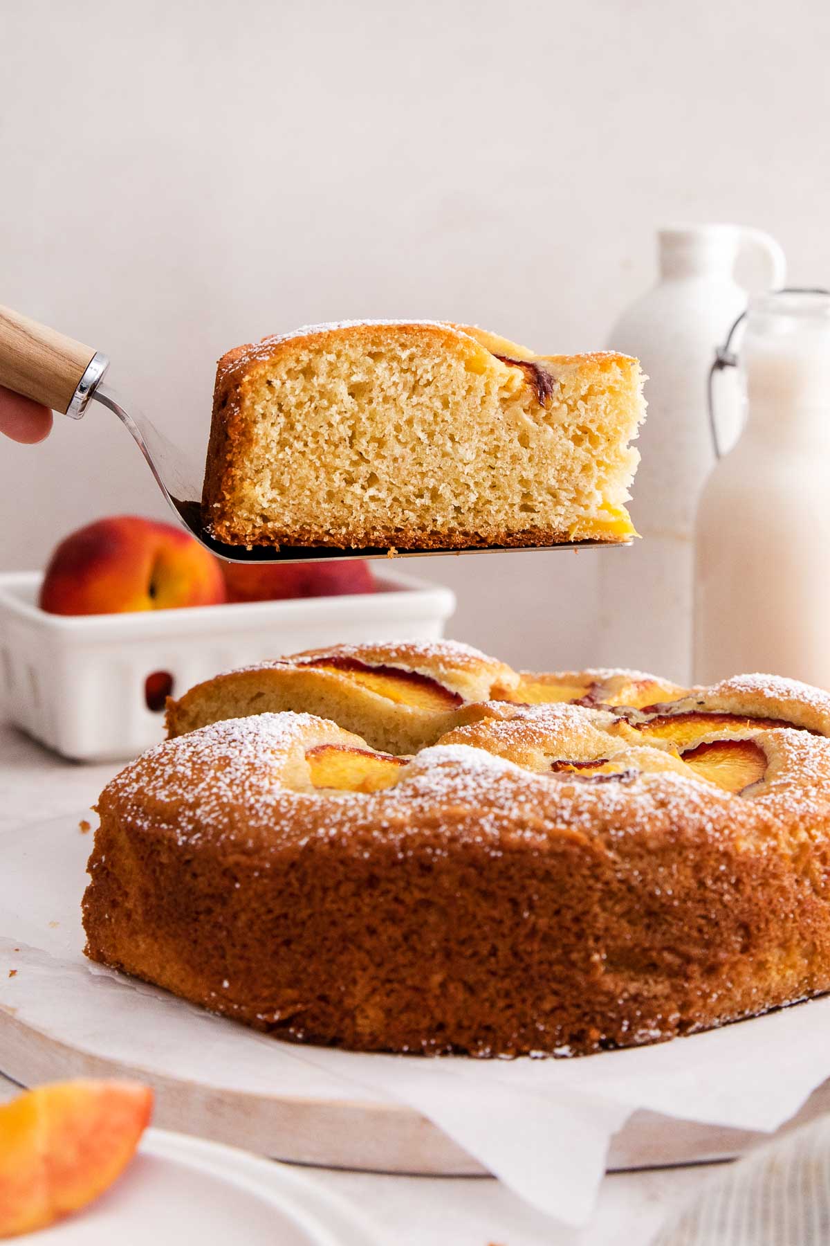 A cake with peach slices and powdered sugar sits on a table. A server lifts a slice, showing its fluffy texture. Peaches and milk bottles are in the background.