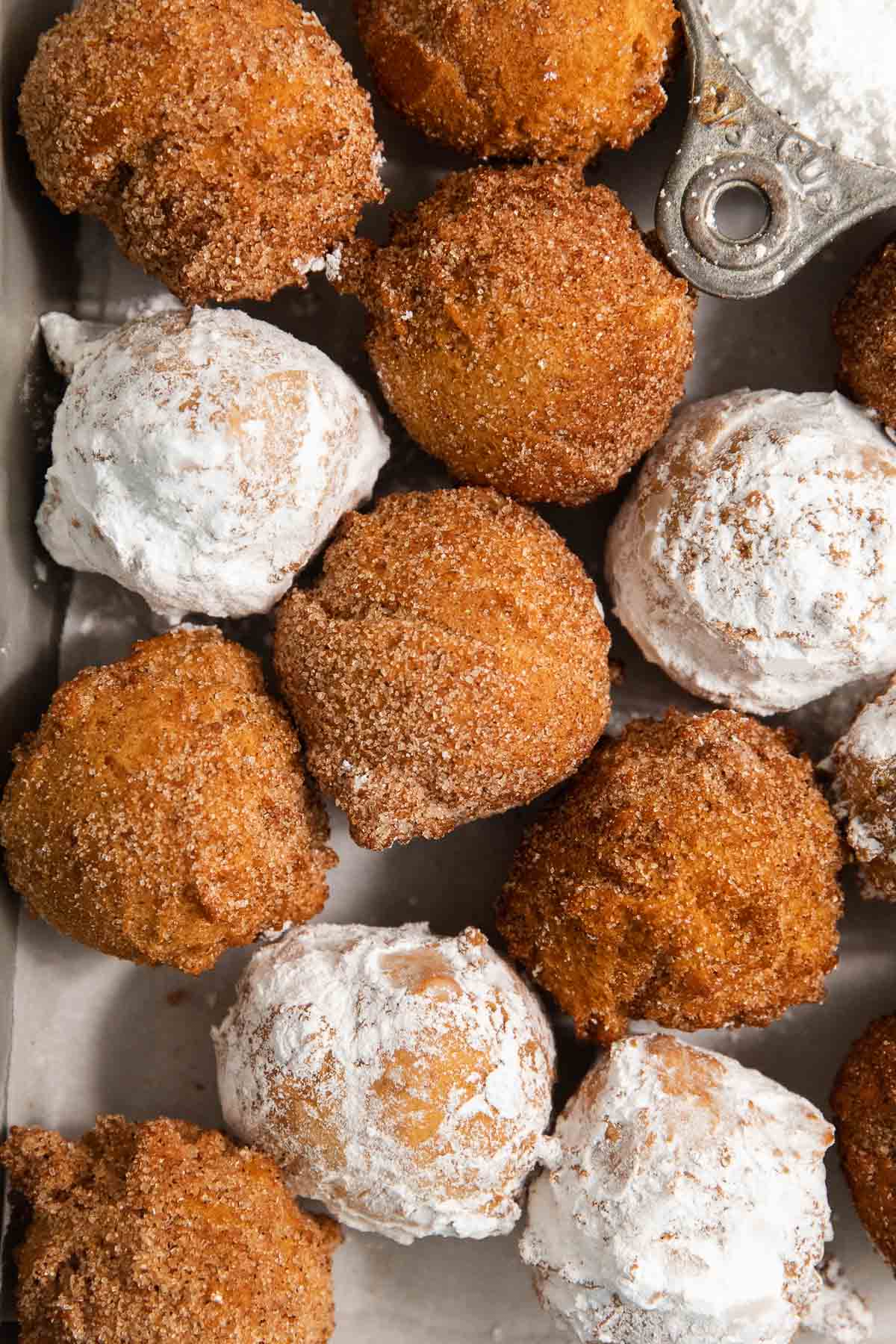 A close-up of several golden-brown and powdered sugar-coated donut holes arranged tightly together on a tray, with a metal scoop holding powdered sugar nearby.