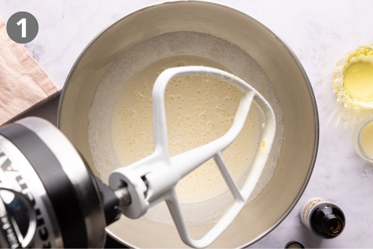 A stand mixer bowl with a paddle attachment beating eggs and sugar; vanilla extract and butter are nearby on a marble countertop. Number 1 is in the top left corner.