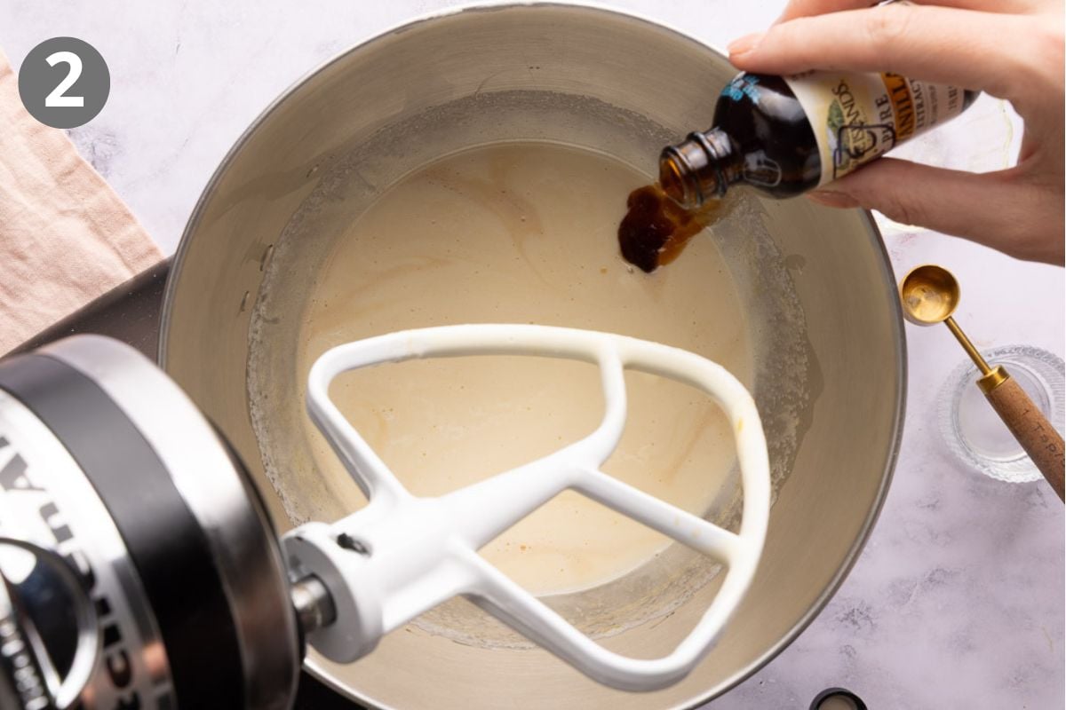 A hand pours vanilla extract into a stand mixer bowl containing a creamy mixture. The mixer has a paddle attachment, and a small bowl with a spoon sits nearby. A number 2 is in the top left corner.