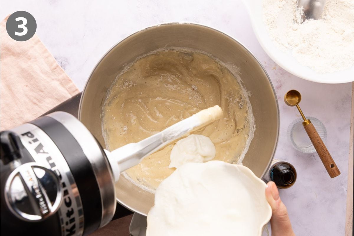 A hand pours yogurt into a mixing bowl with batter, attached to a stand mixer. A bowl of flour, measuring spoon, and vanilla extract are nearby. The image is labeled with the number 3 in the top left corner.