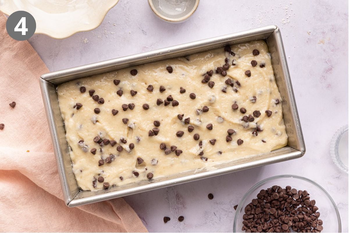 A loaf pan filled with unbaked batter topped with chocolate chips sits on a light surface, surrounded by a pink cloth, a bowl of mini chocolate chips, and a small empty dish.