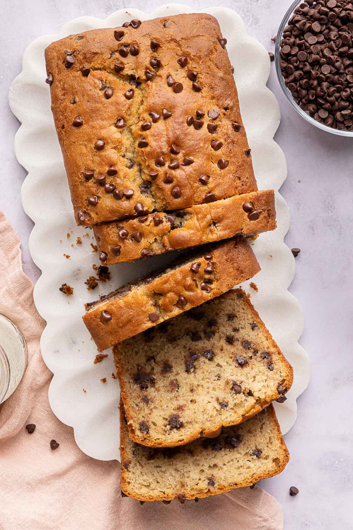 A chocolate chip yogurt loaf on a white platter, partially sliced, showing moist texture and chocolate chips inside. A bowl of chocolate chips and a pink cloth are nearby on a light surface.