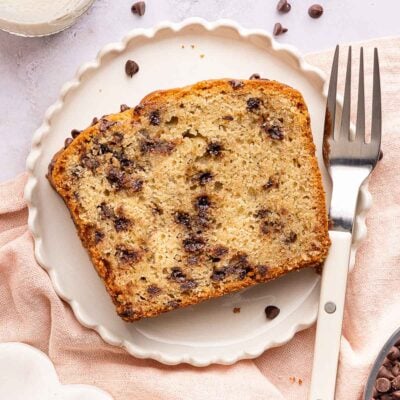 A slice of chocolate chip yogurt cake on a white plate with a fork beside it. Chocolate chips are scattered around, and a pink napkin is partially visible underneath the plate.