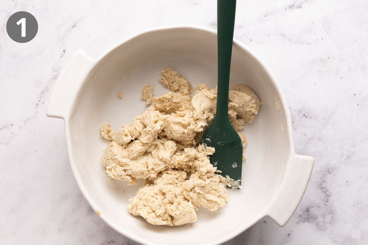 White bowl with rough cookie dough and a dark green spatula on a white marble surface. The number 1 is in the top left corner, indicating the first step in a recipe.