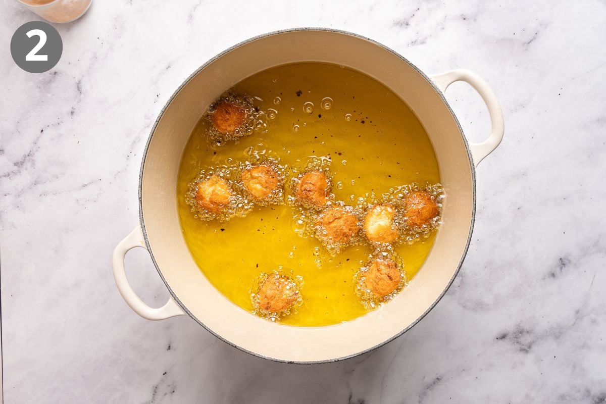 Eight golden-brown balls are frying in hot oil inside a round, white pot on a light marble surface. A small 2 in a gray circle is in the top left corner.