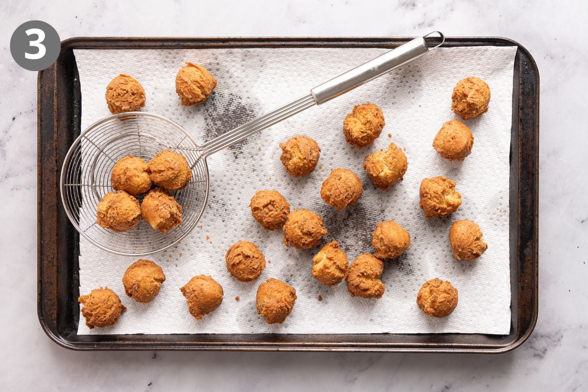 A baking tray with fried golden-brown balls on paper towels. A metal skimmer holds some balls. A circle with the number 3 is in the top left corner.