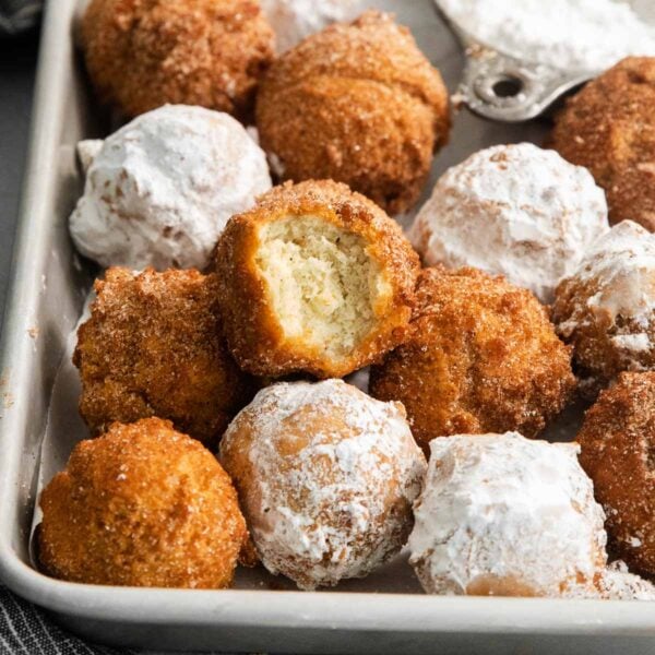 A tray of donut holes, some coated in powdered sugar and others in cinnamon sugar. One donut hole with a bite taken out is stacked on top, showing the soft inside.