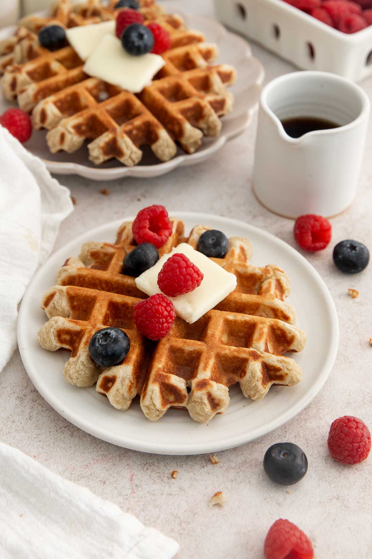 A plate with a waffle topped with butter, raspberries, and blueberries. Syrup in a small pitcher and more berries are nearby. Another waffle is partially visible in the background.