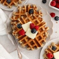 Two waffles on a plate topped with butter, blueberries, and raspberries. A fork rests on the plate, and extra berries are scattered around on the table.