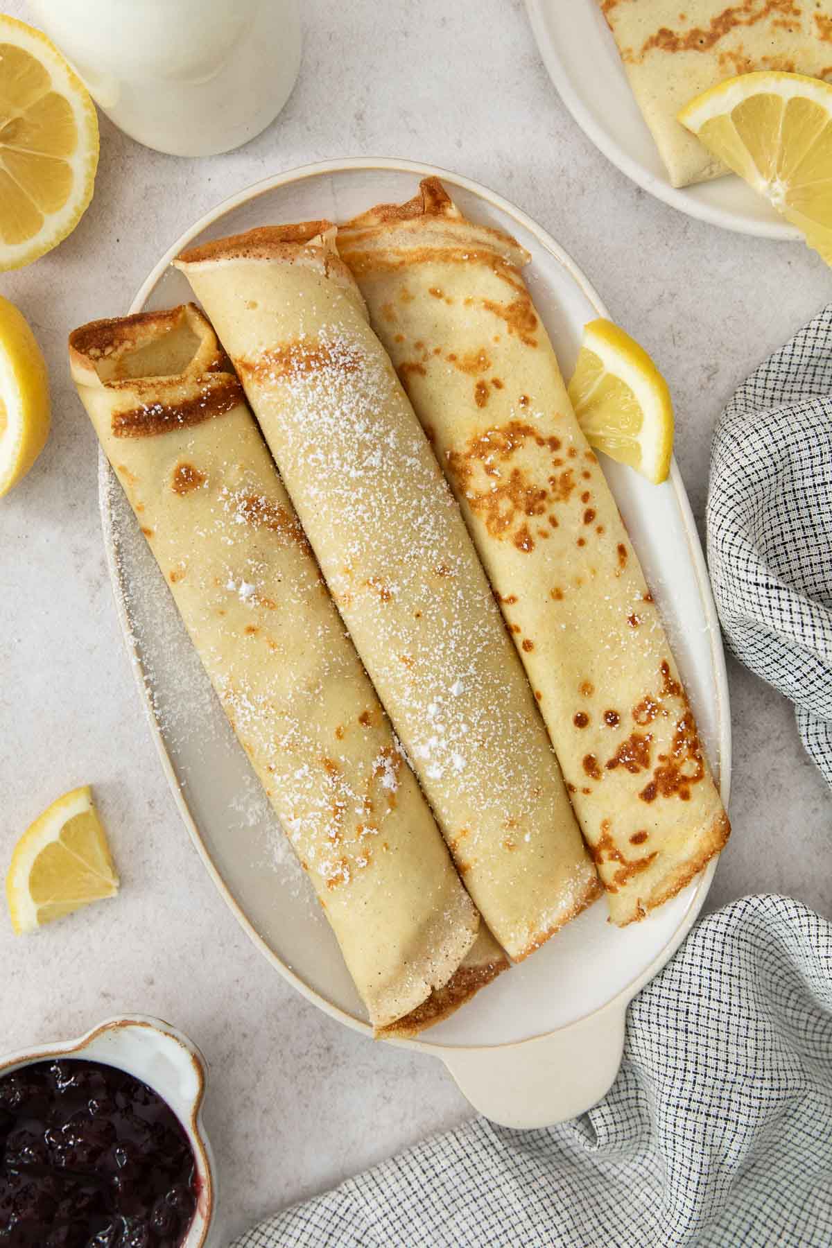 Three rolled swedish pancakes on a white plate, lightly dusted with powdered sugar, served with lemon wedges. A checkered napkin, a partial plate, and a small bowl of dark jam are nearby.
