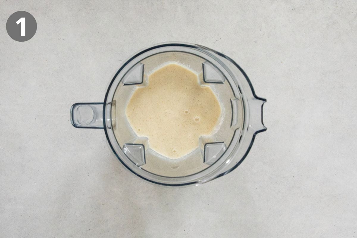 Top view of a blender with a creamy, light beige mixture inside, placed on a light-colored surface. A gray circle with the number 1 is in the upper left corner.