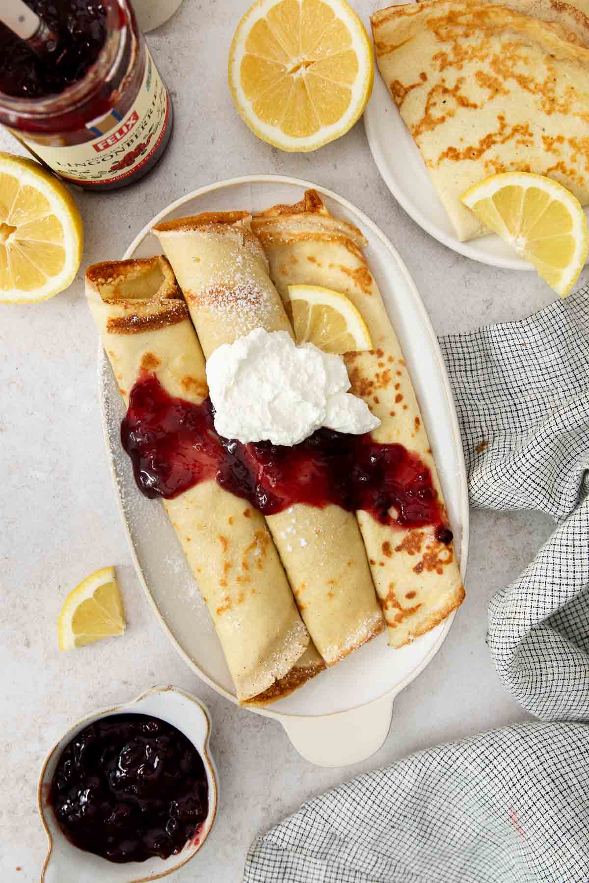 Three rolled swedish pancakes topped with berry jam and whipped cream on a white plate, surrounded by lemon slices, a jar of jam, and a small bowl of jam on a light surface.